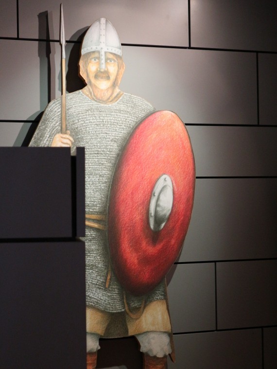 Das Foto zeigt eine lebensgroße Holzfigur mit der Zeichnung eines Ritters darauf. Er trägt in der rechten Hand einen Speer, eine Helm mit Nasal, ein Kettenhemd und in der Linken einen Schild.