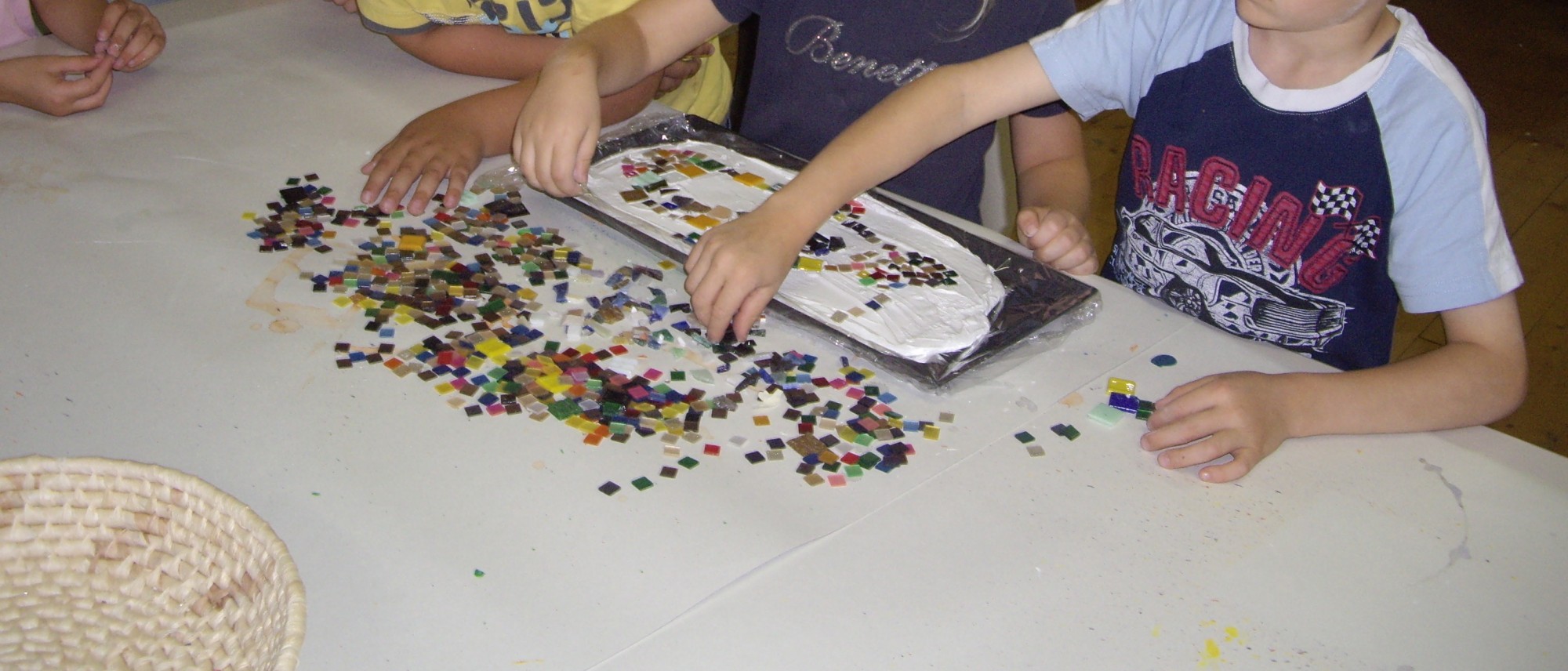 Das Foto zeigt Arme und Hände von Kindern beim Legen eines kleinen Mosaikes. Sie greifen nach den bunten Steinchen und verteilen sie auf eine weiße Paste in einer länglichen Unterlage.