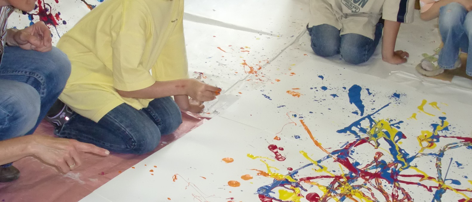 Das Foto zeigt Kinder und eine erwachsene Person, die sich hockend und kniend um ein großes, mit bunten Farben bespritztes Papier gruppieren. Die Gesichter der Personen sind nicht zu sehen.