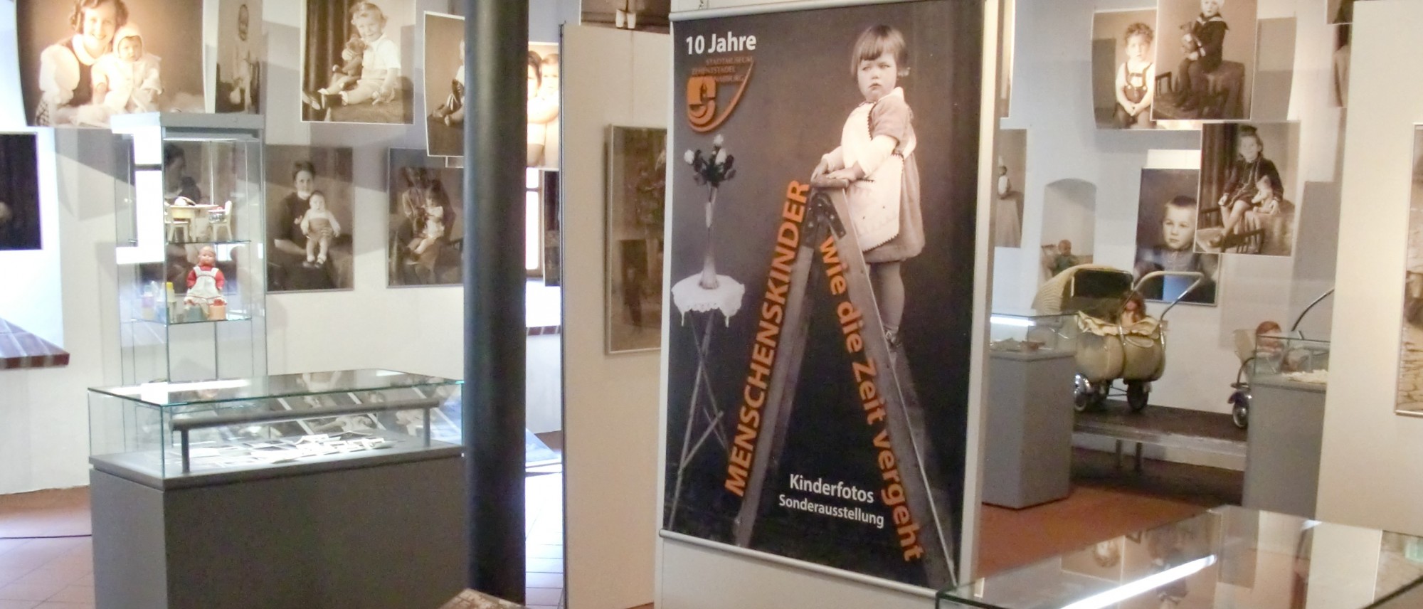 Das Foto zeigt einen Ausschnitt der Ausstellung &quot;Menschenskinder&quot;. Im Vordergrund ist ein großes Plakat mit einem kleinen Mädchen auf einer Leiter zu sehen. Es hängt an einer Stellwand. Im Hintergrund hängen große Ausdrucke von älteren Porträtfotos mit Müttern und ihren Kindern. 