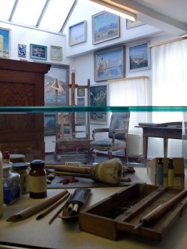 Das Foto zeigt einen Ausschnitt des Ateliers von Karl Schmidt. Im Vordergrund liegen Malutensilien wie Pinsel und Farbe in einer Vitrine. Im Hintergrund eine Staffelei und anderes Mobiliar.